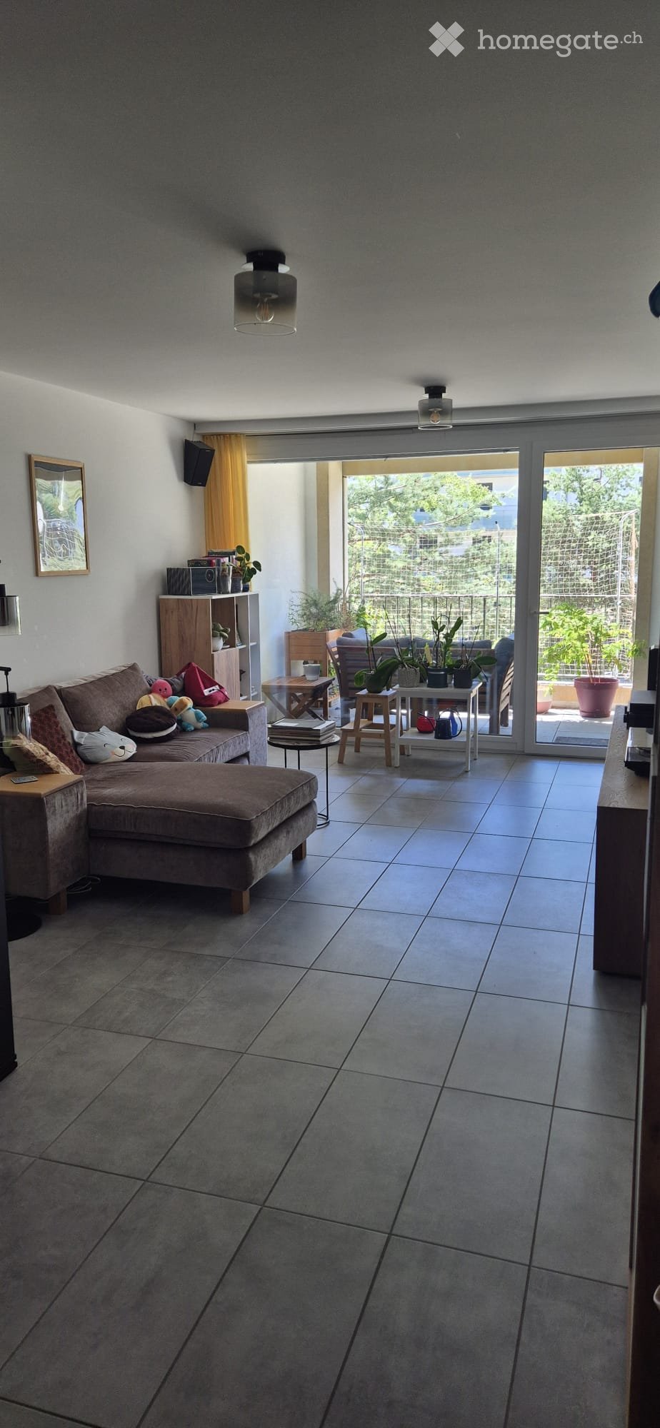 Modern living room with grey sofa, ottoman, small tables, and shelves. Large sliding glass doors open to a balcony with plants.