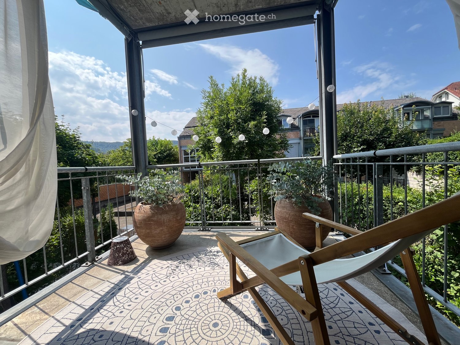 Balcony with a wooden folding chair, two large potted plants, and a decorative small pot. The balcony has a metal railing and a patterned tile floor.