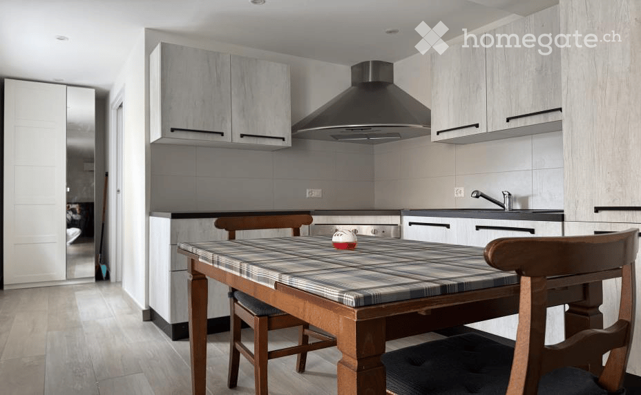 Modern kitchen with light grey cabinets, a central dining table with chairs, and a stainless steel range hood above the stove.