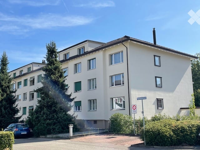 A multi-story residential building with white walls, multiple windows, and green shutters. There is a parked car in front and some greenery around the building.