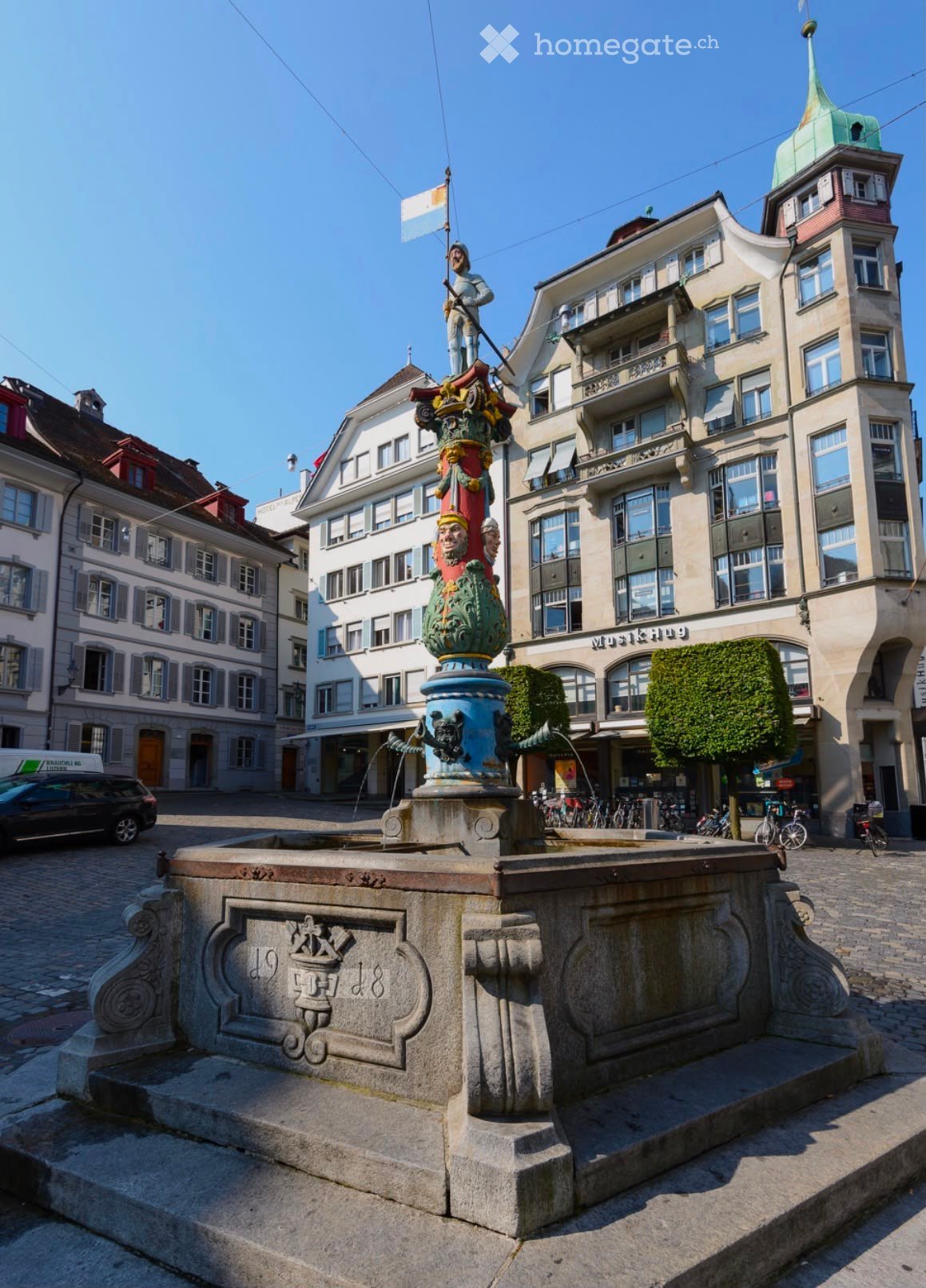 A historic square with a decorative fountain, surrounded by multi-story buildings with classic European architecture, bicycles, and a parked car.