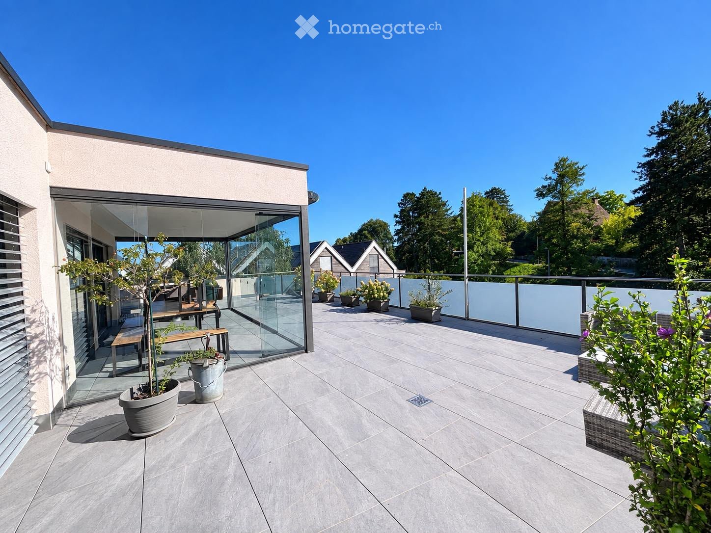 Modern balcony with grey tiled flooring, glass railing, and potted plants, featuring a small bench and a view of surrounding greenery.