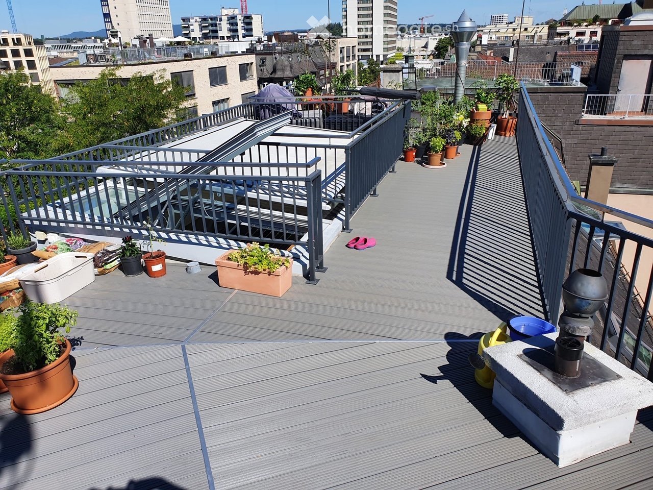 Rooftop terrace with wooden decking, metal railings, potted plants, and a view of the city skyline.
