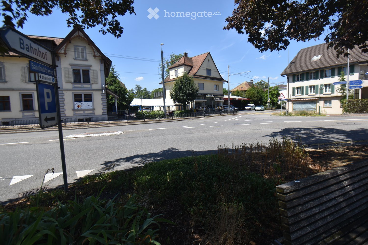 A street scene with several buildings, including a train station sign, a parking sign, and a small shop with a canopy. The road is empty, and there are some trees and greenery visible.