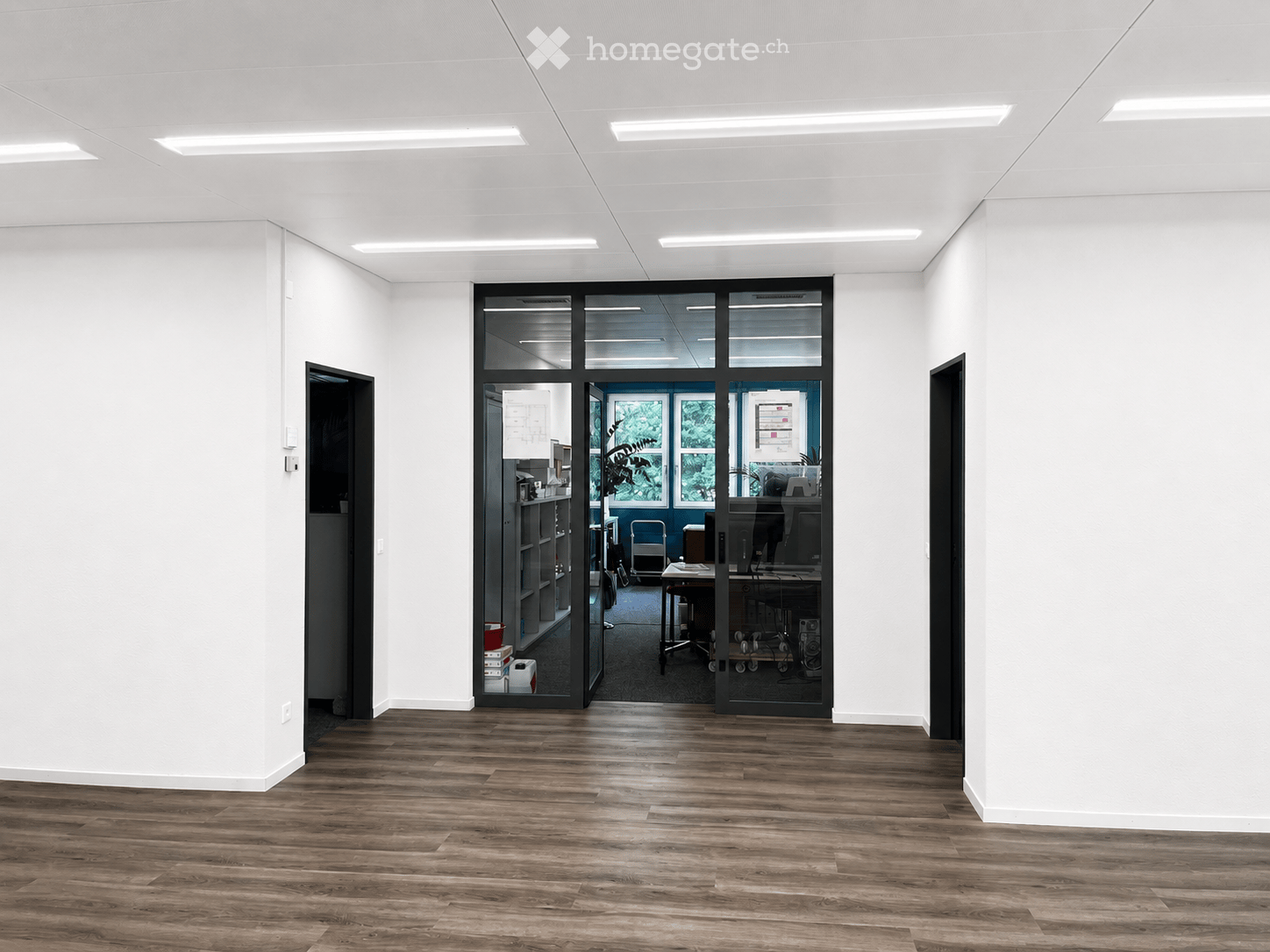 Empty room with wooden flooring, white walls, and recessed lighting. A glass door leads to an adjacent office space with desks, chairs, and office equipment.