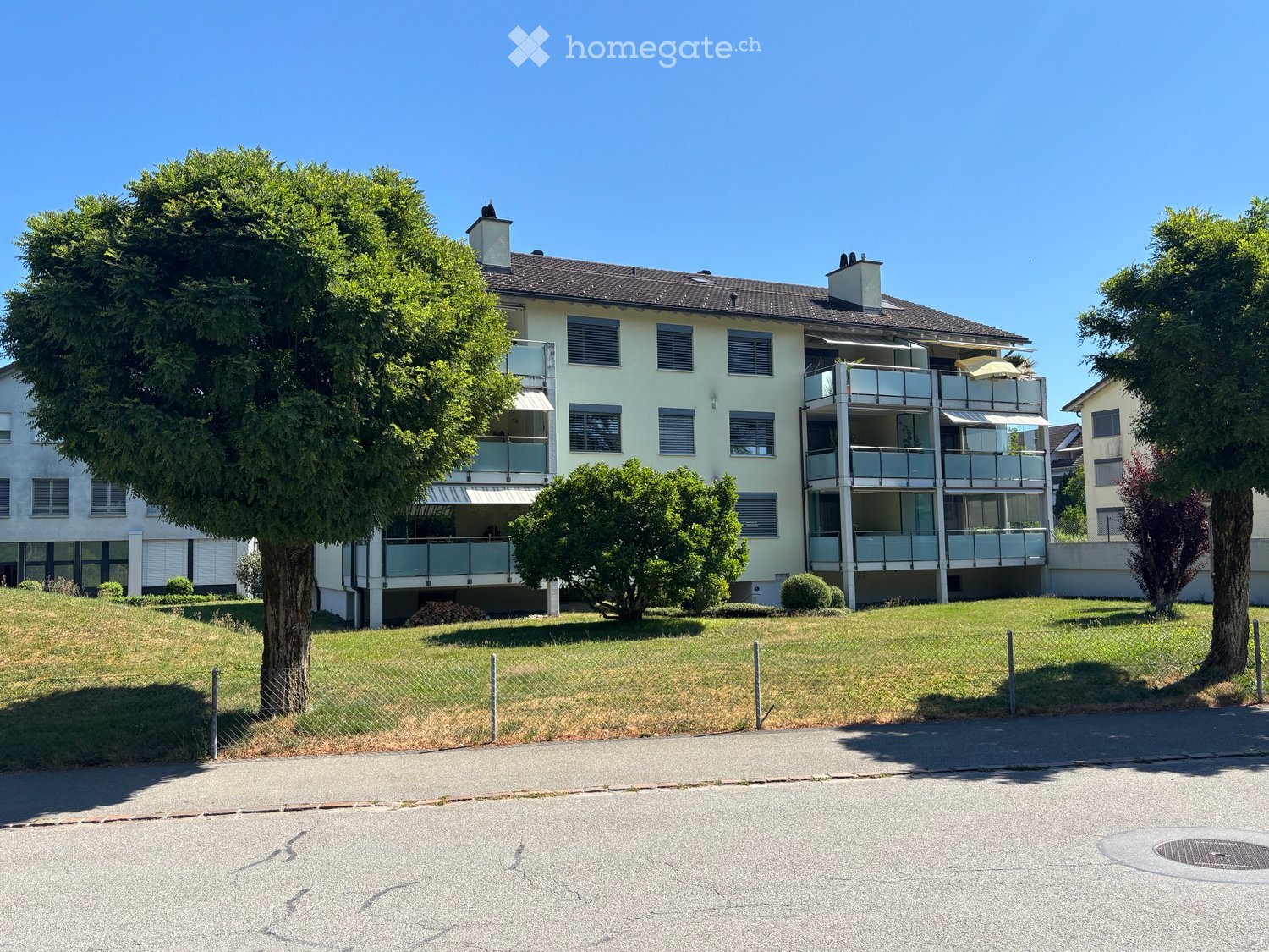 A multi-story residential building with balconies, surrounded by greenery and a fenced lawn area.