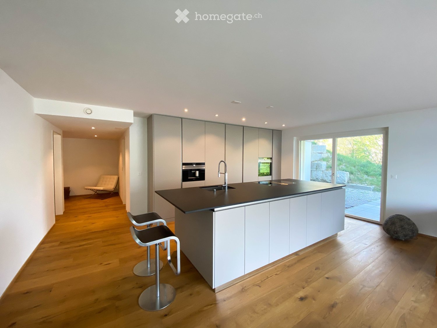 Modern kitchen with white cabinets, dark countertop, island, two stools, and a sink with a modern faucet. The space features wooden flooring and large windows with a view of greenery.