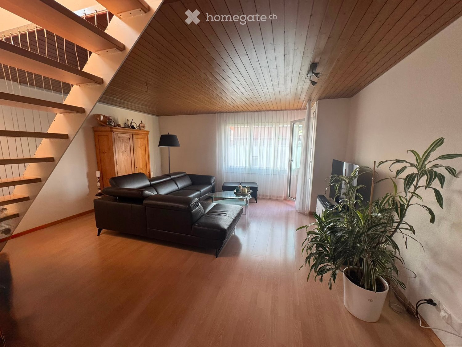 Modern living room with wooden flooring, a black leather sofa, a glass coffee table, a floor lamp, a television, and a large potted plant. A staircase leads to an upper level.