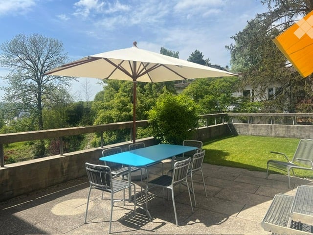 Balcony with a large umbrella, blue table, and chairs, overlooking a green lawn and trees.