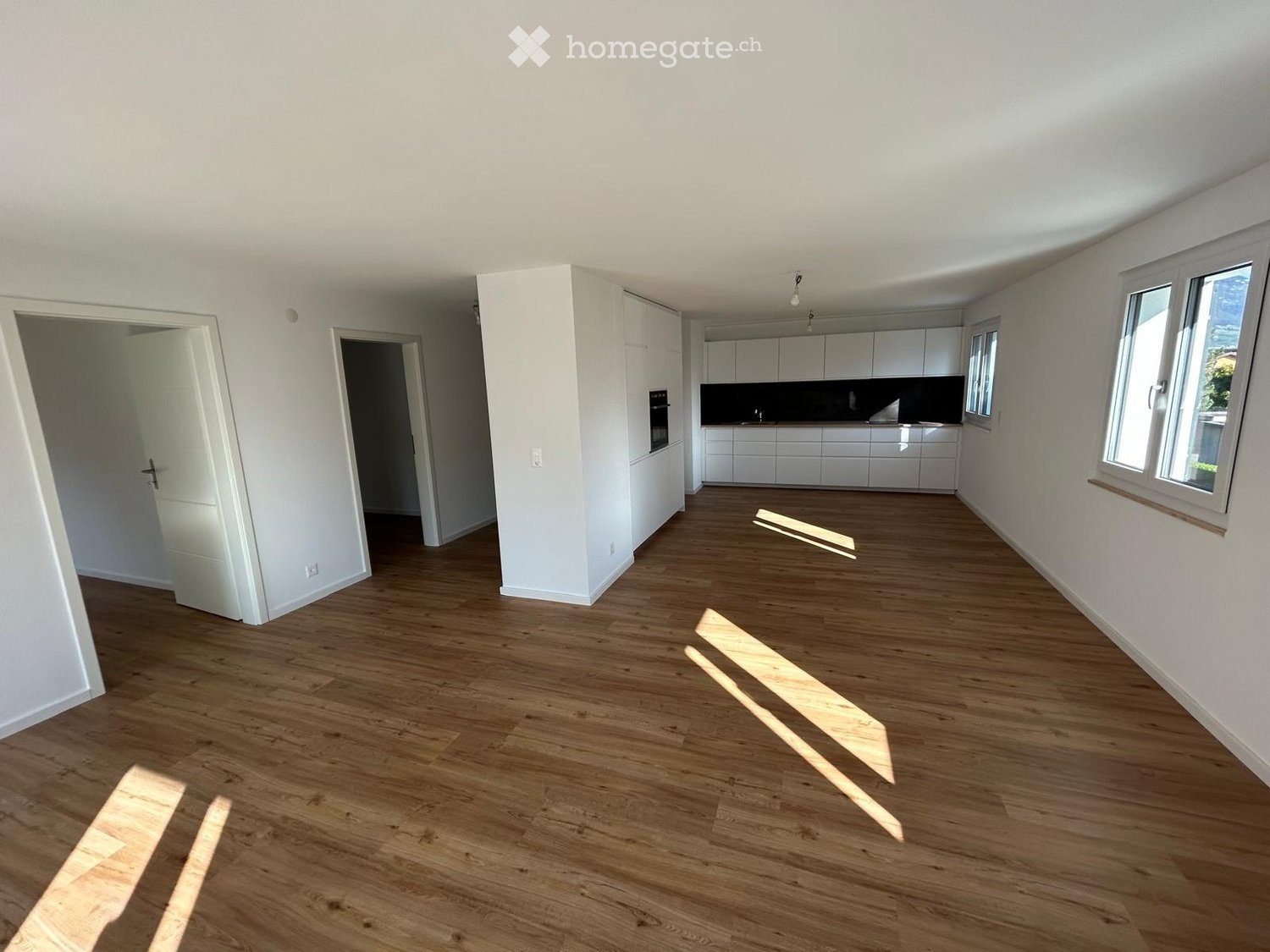 Empty room with wooden flooring, white walls, and large windows. The adjacent kitchen area features white cabinets and a black countertop.
