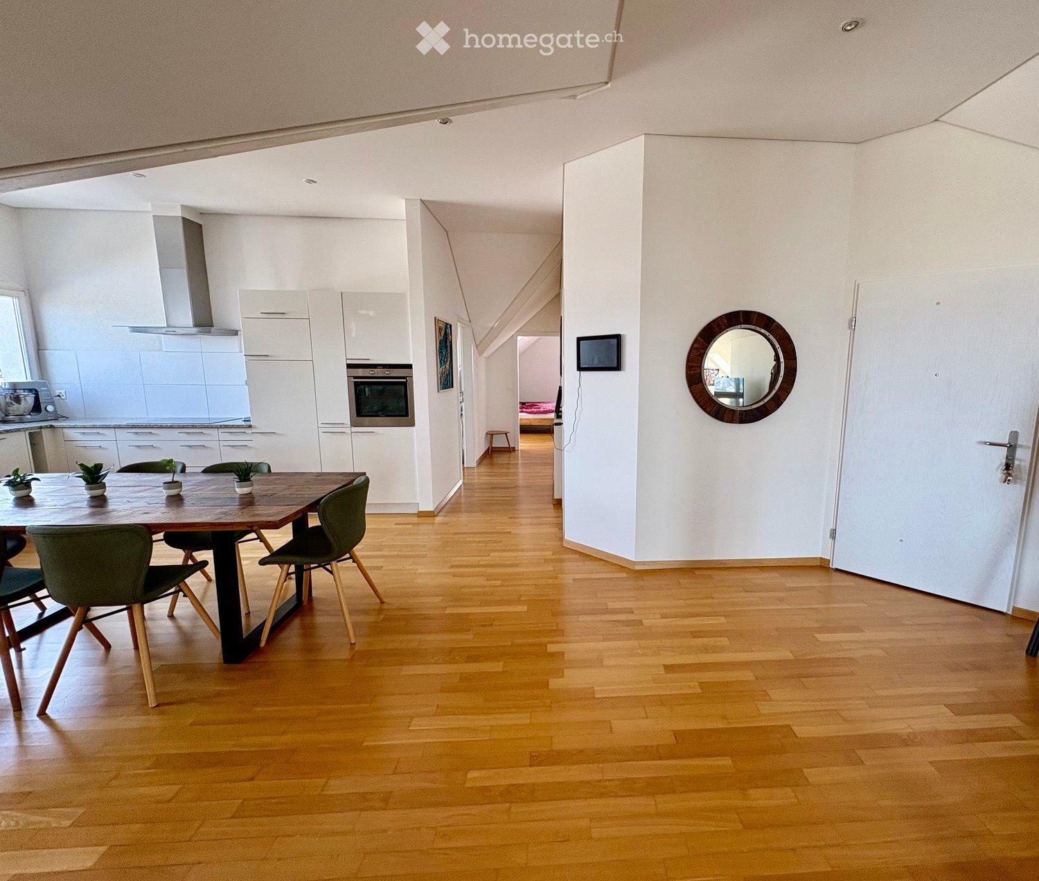 Bright, modern dining area with wooden flooring, a wooden dining table surrounded by green chairs, a round wall mirror, and a small screen mounted on the wall. The area connects to a modern kitchen with white cabinets and stainless steel appliances.
