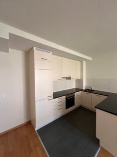 Modern kitchen with white cabinets, black countertops, and a built-in oven. The space features wooden flooring and ample natural light from large windows.