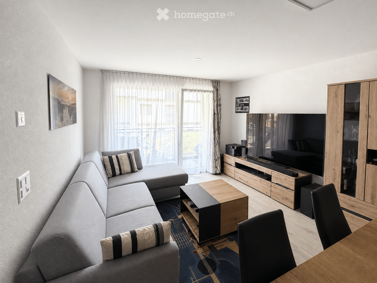 Modern living room with a grey sectional sofa, wooden coffee table, TV stand, and a dining area with a wooden table and chairs. Large windows with sheer curtains offer a view of the balcony.