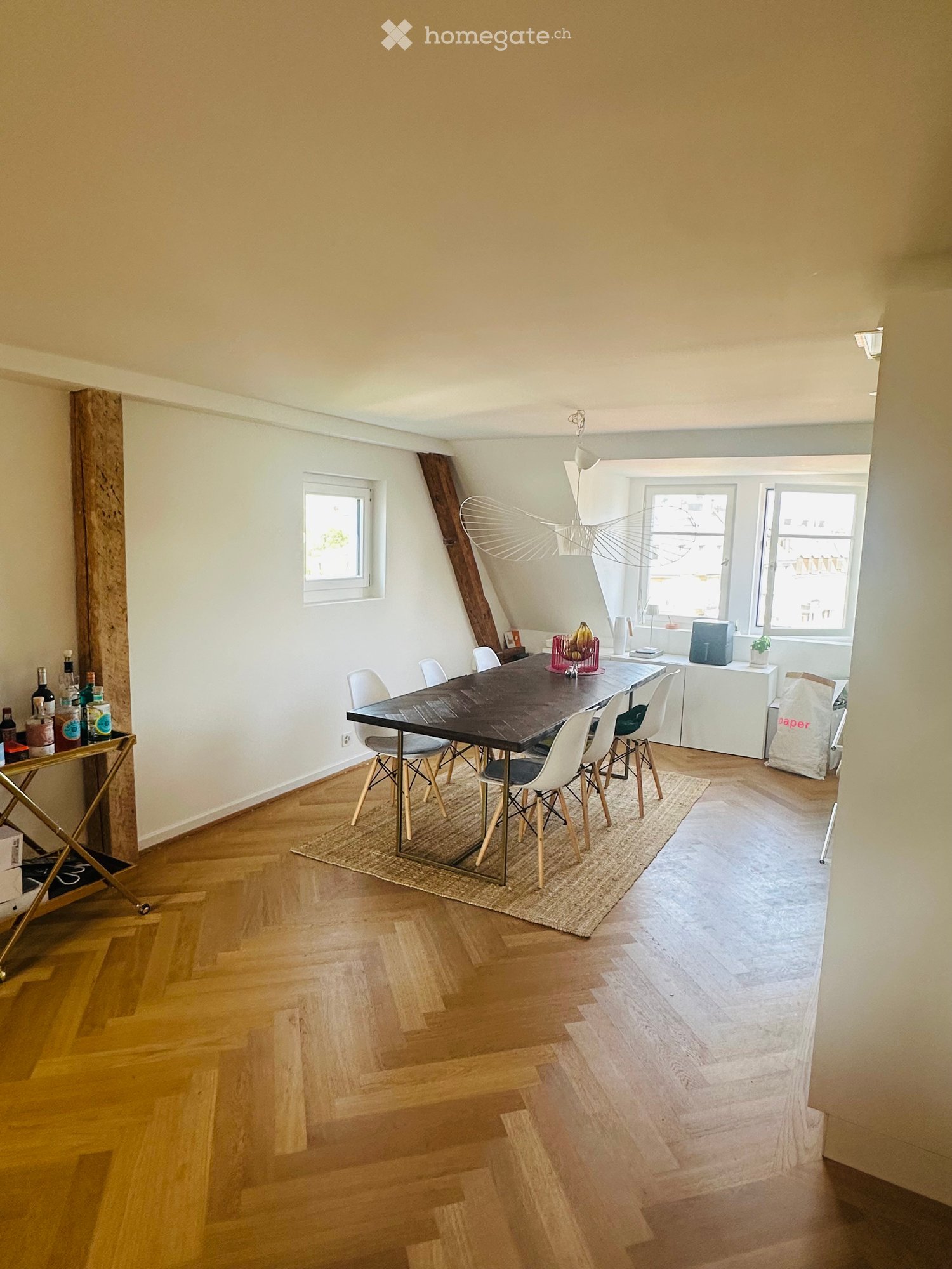 Dining room with wooden floor, large dining table surrounded by chairs, small bar cart with bottles, and a view of a kitchen area with a refrigerator and plant.