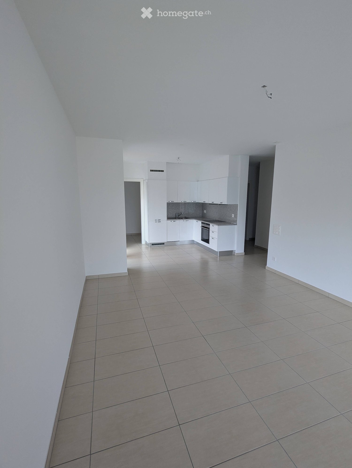 Empty room with tiled floor and white walls, featuring a small kitchen area with white cabinets and appliances.