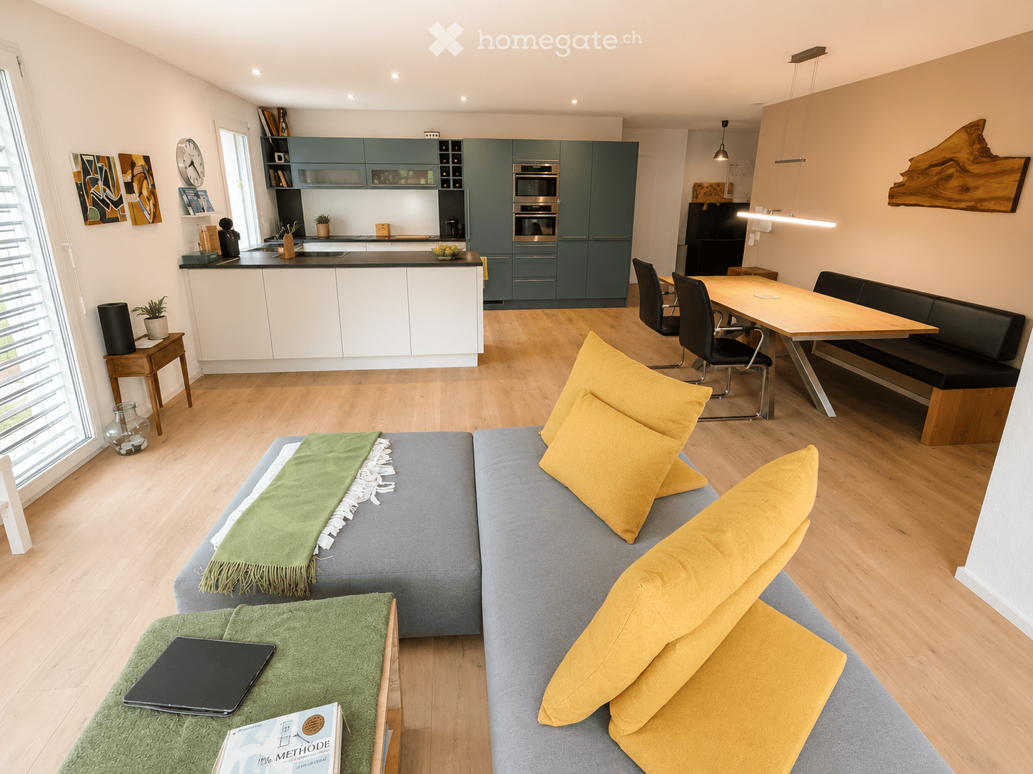 Open-plan living and dining area with a grey sofa, yellow pillows, coffee table, laptop, books, and a modern kitchen with green cabinets and stainless steel appliances.