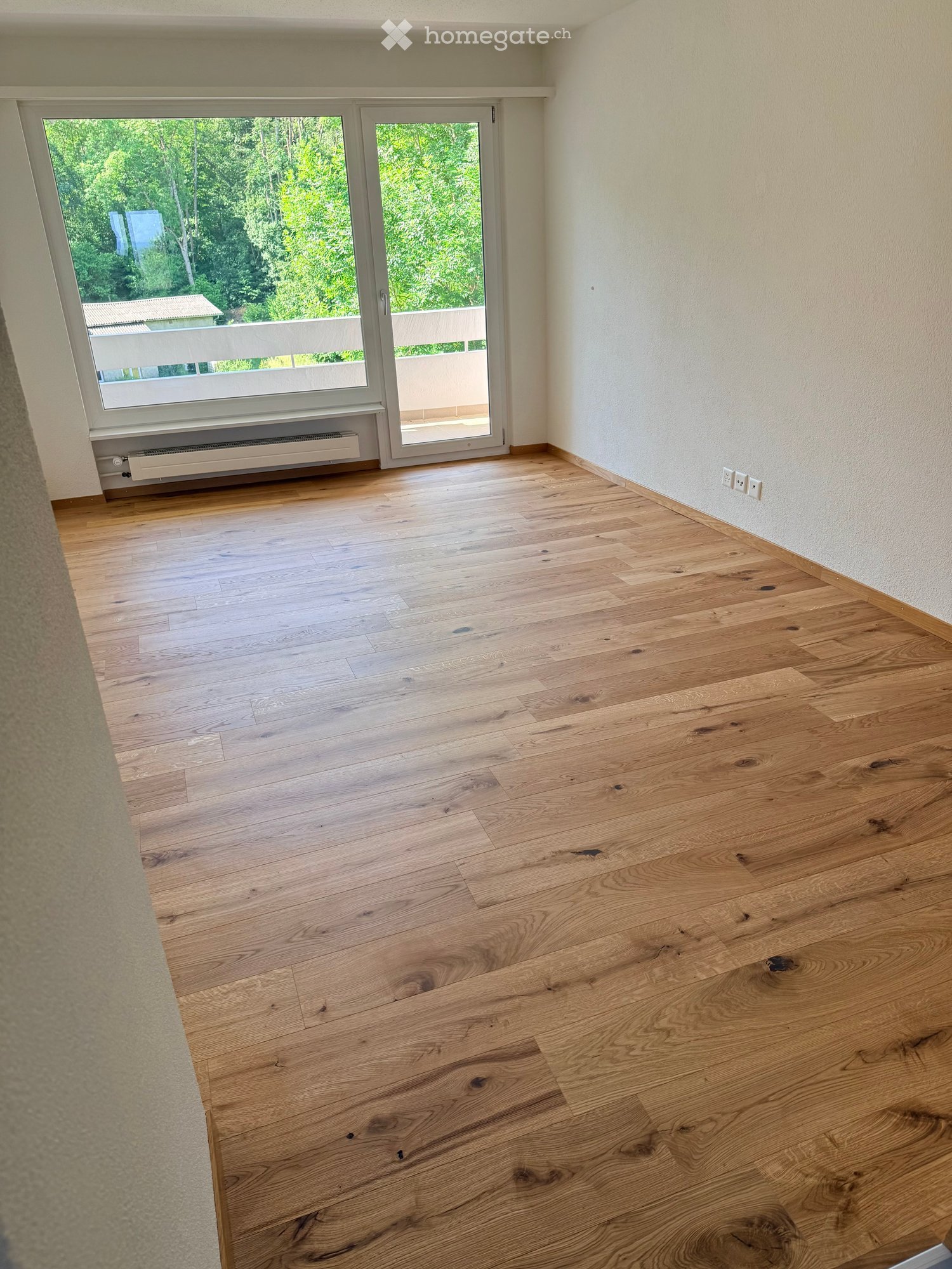 Empty room with light wooden flooring, white walls, large window and sliding glass door leading to a balcony.
