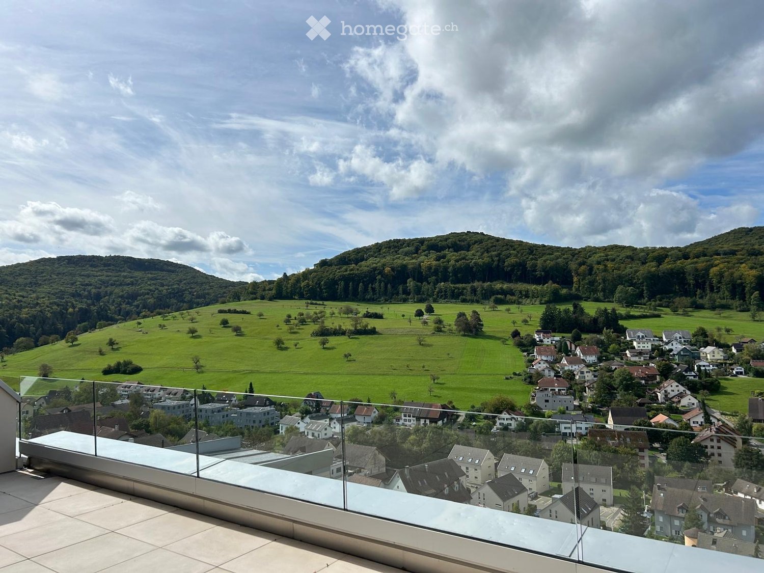 Balcony with a panoramic view of a green valley, scattered houses, and distant hills under a partly cloudy sky.