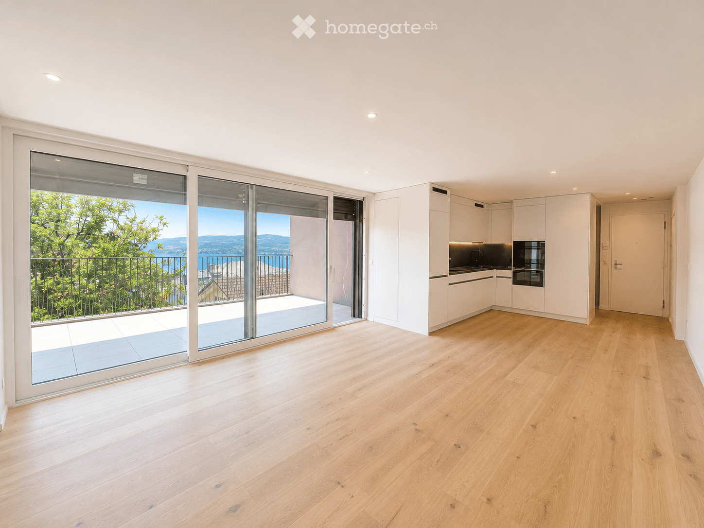 Bright, open-plan space with light wooden flooring, large sliding glass doors leading to a balcony with scenic views, and a modern white kitchen area with cabinets, sink, and oven.