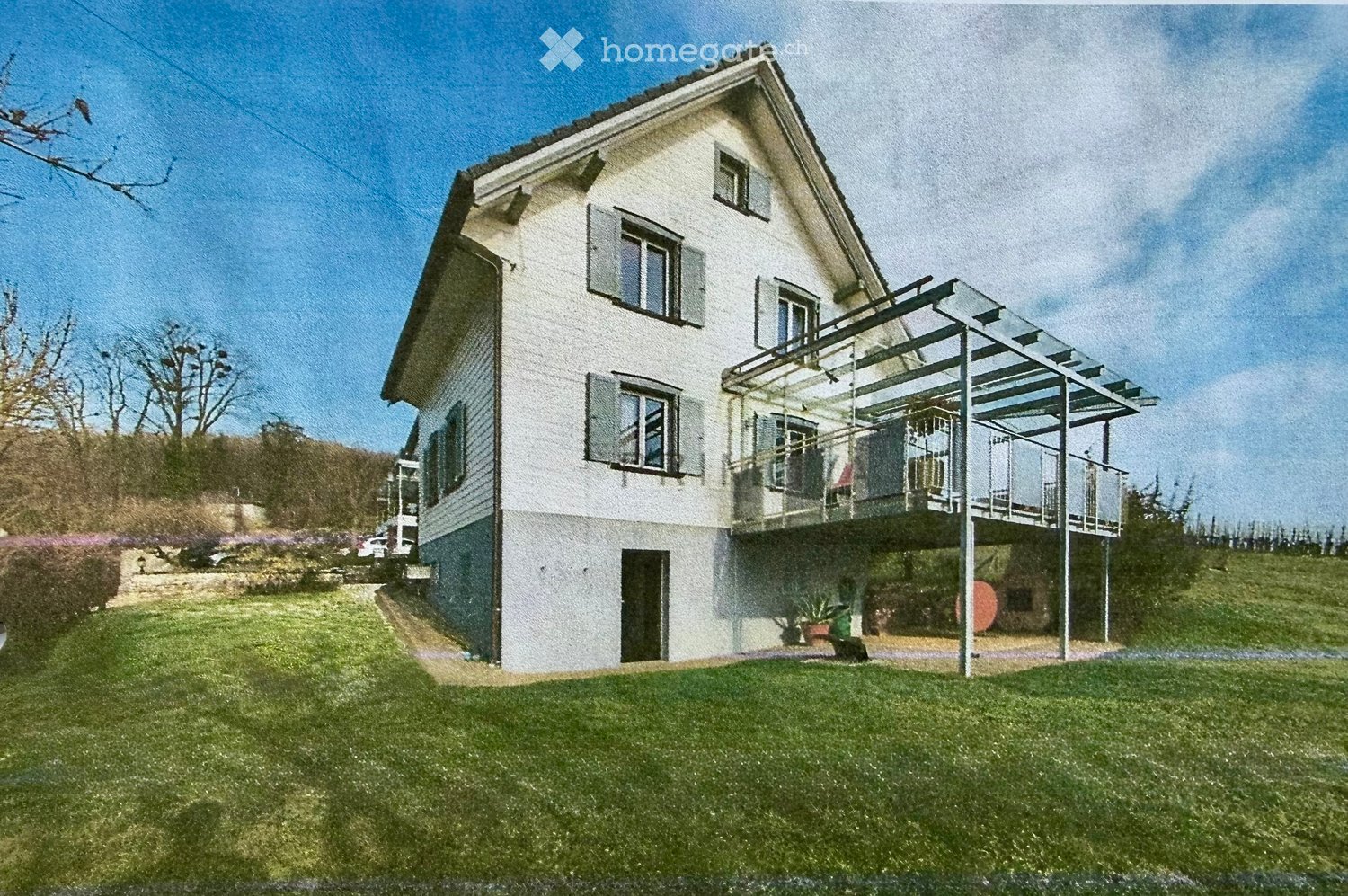 Two-story house with white exterior, multiple windows, and a covered balcony on the upper level. The house is surrounded by a well-maintained lawn and garden area.