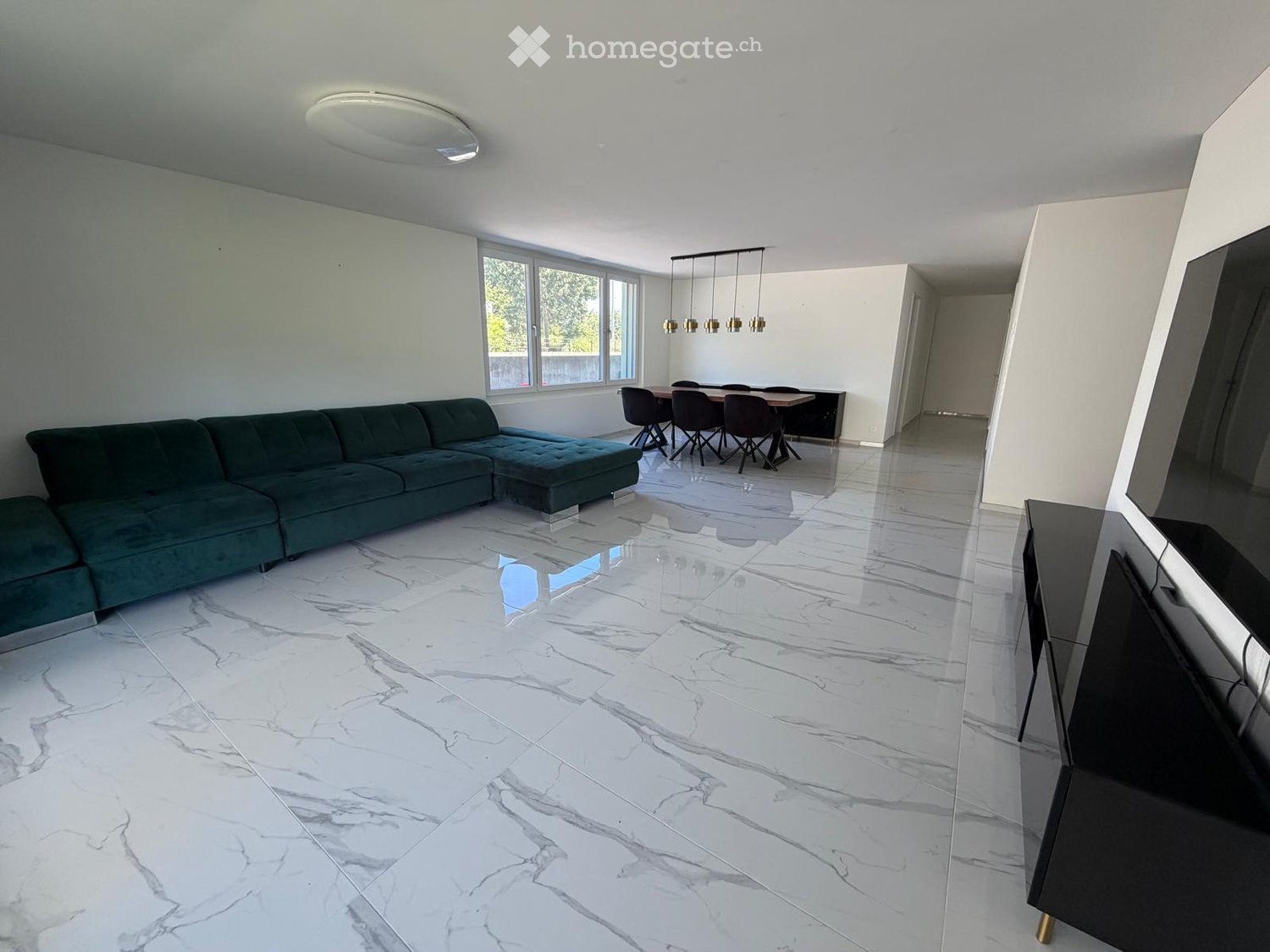Open-plan living and dining area with a green sectional sofa, ottoman, dining table with chairs, and a television stand. The room features a polished marble floor and large windows.