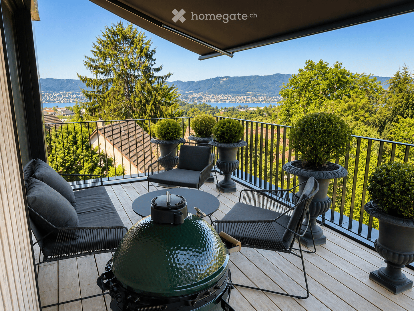 Balcony with wooden decking, black metal railing, and scenic mountain and lake view. Features a green grill, black sofa, chairs, and a small table.