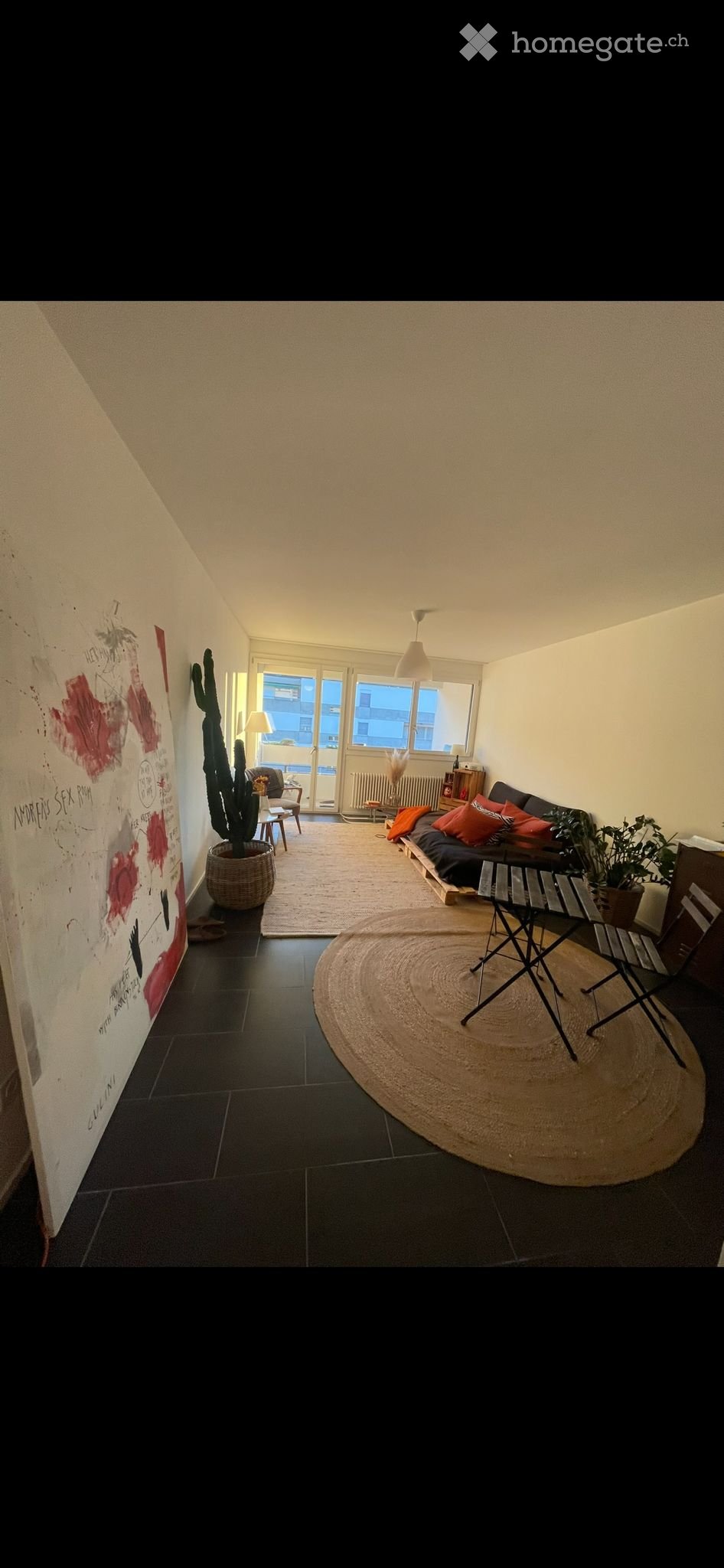 A modern living room with a dark tiled floor, a large round jute rug, a sofa with colorful cushions, a potted cactus, a small table, and a hanging lamp.