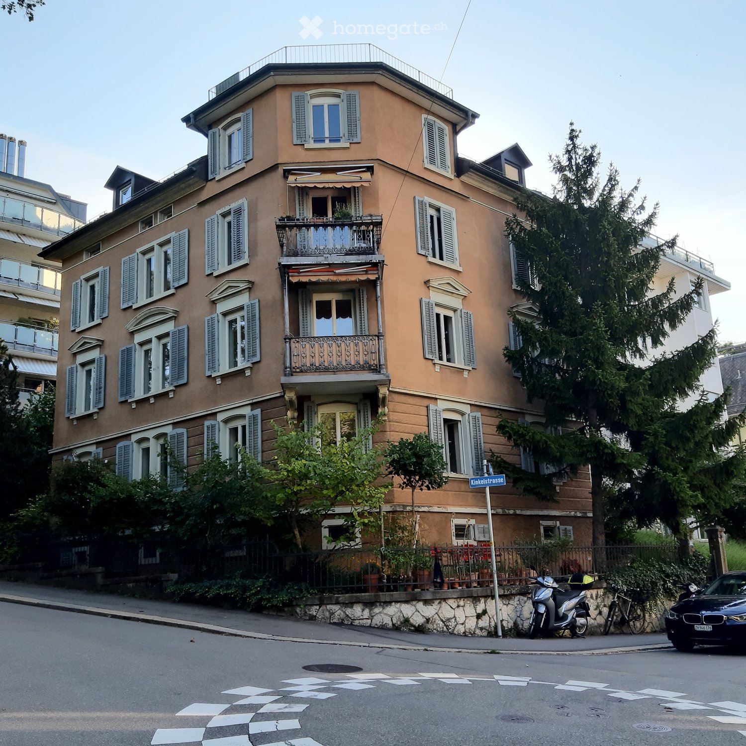 A multi-story residential building with beige facade, multiple windows with shutters, balconies, and decorative railings. The building is surrounded by greenery and parked vehicles.
