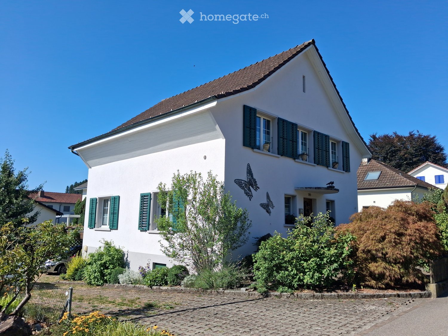 Two-story white house with green shutters, butterfly wall art, and a well-maintained garden with shrubs and flowers.