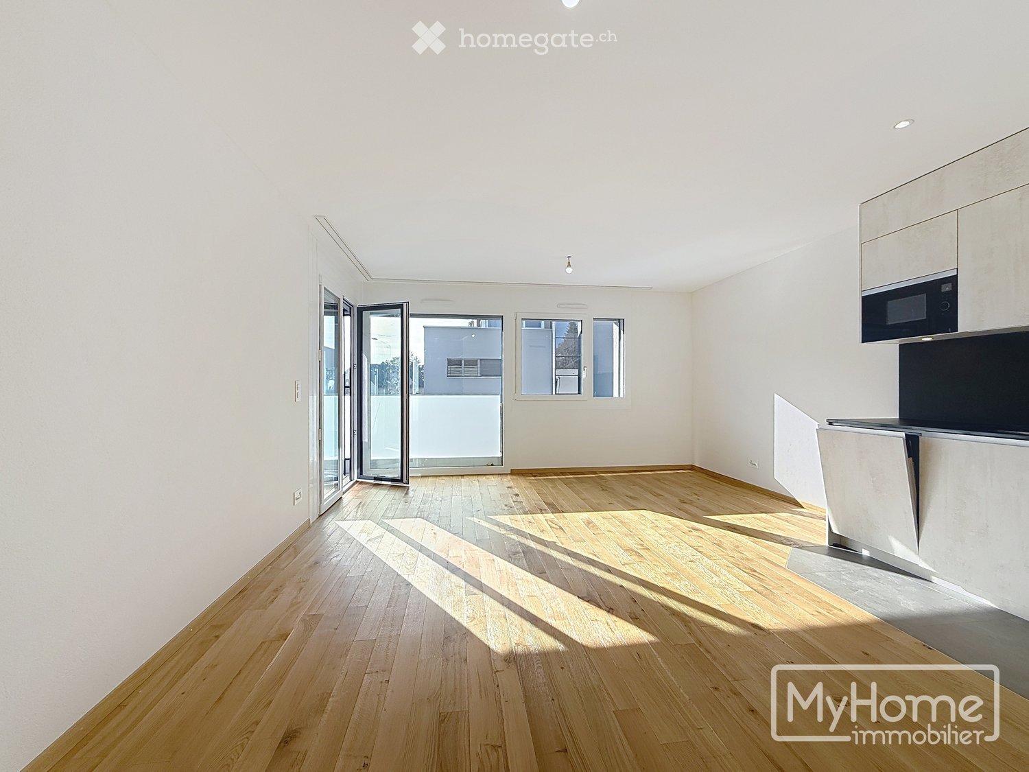 Empty room with light wooden flooring, white walls, and large windows allowing natural light. Features a modern kitchen area with cabinets and a microwave, and a sliding glass door leading to a balcony.