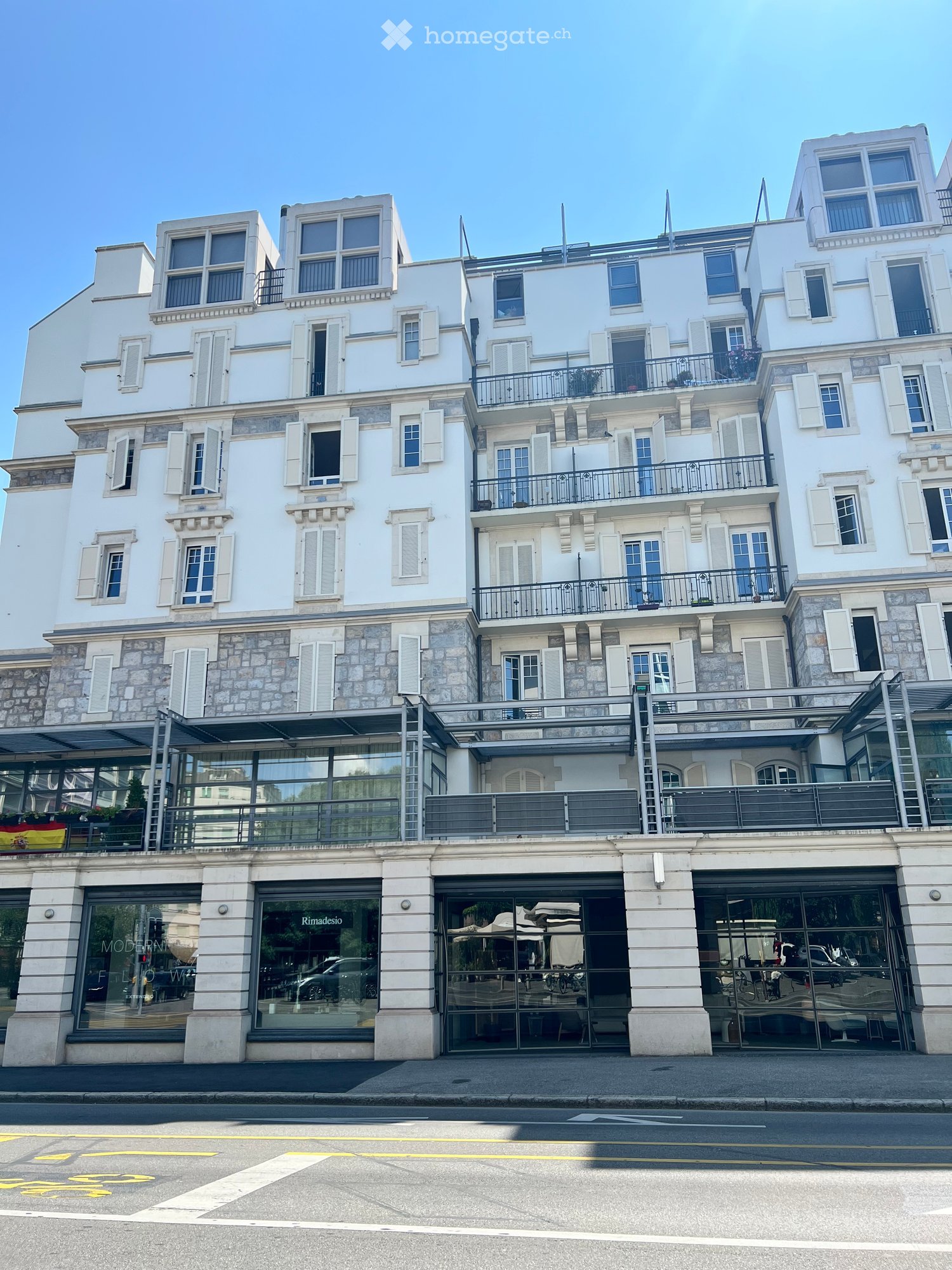 A multi-story white building with stone accents, large windows, and balconies. The ground floor features glass storefronts with signage for "Rimadesio" and "MODERNISMO".