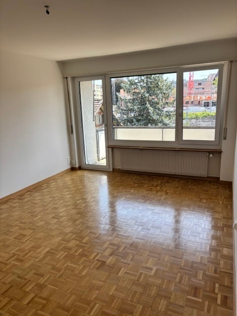 Empty room with wooden parquet flooring, large windows, and a sliding glass door leading to a balcony.