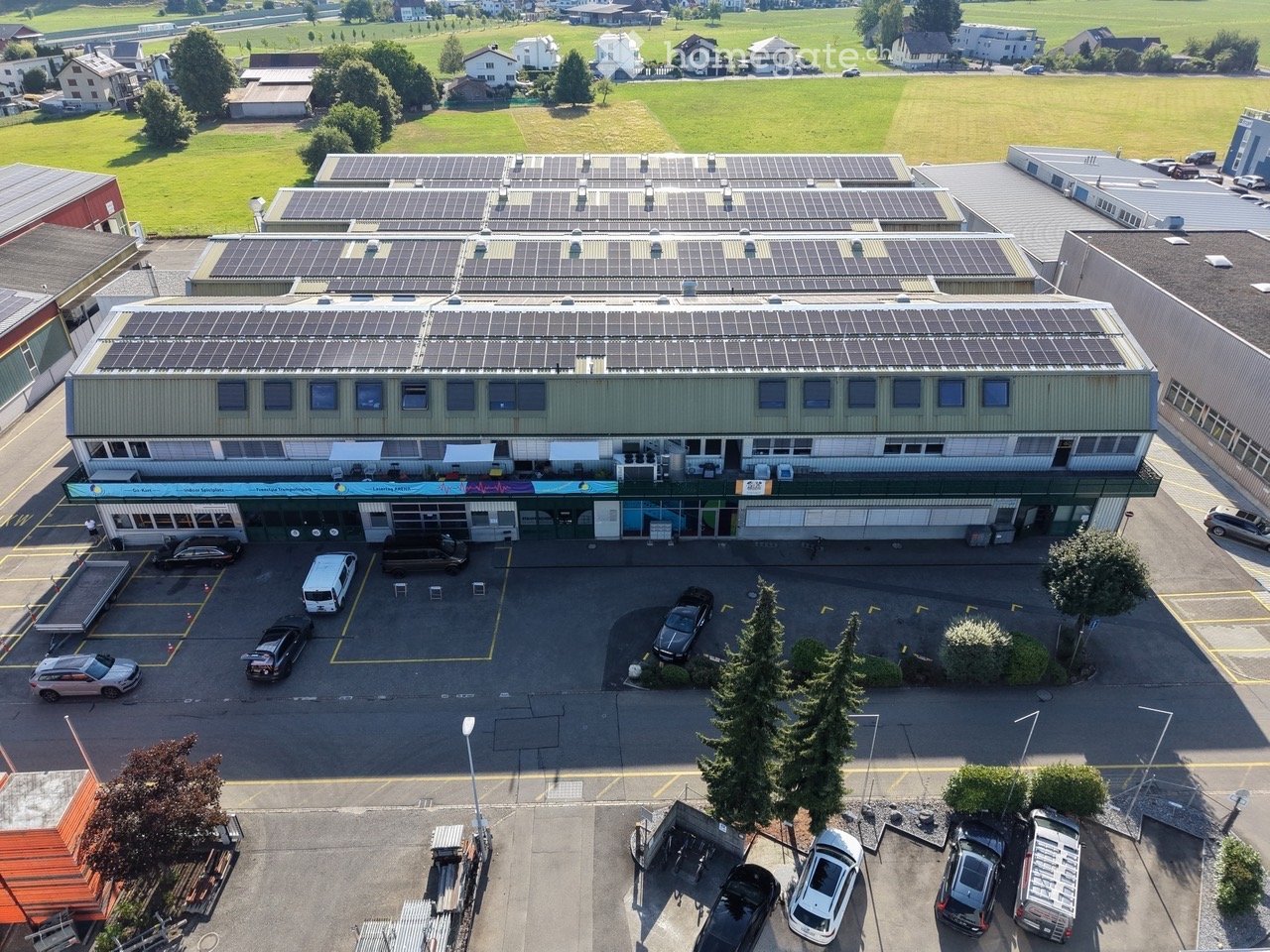 Aerial view of a two-story building with solar panels on the roof, surrounded by a parking lot with several cars and a small garden area.