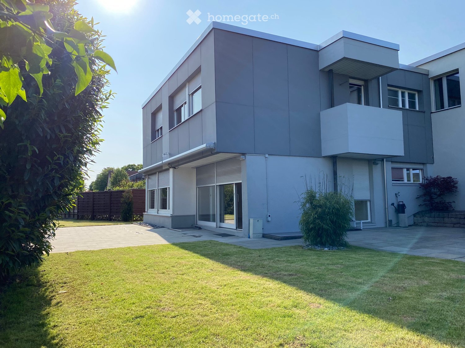 Modern two-story house with grey and white exterior, large windows, and a balcony on the upper level. The house is surrounded by a well-maintained lawn and paved pathways.