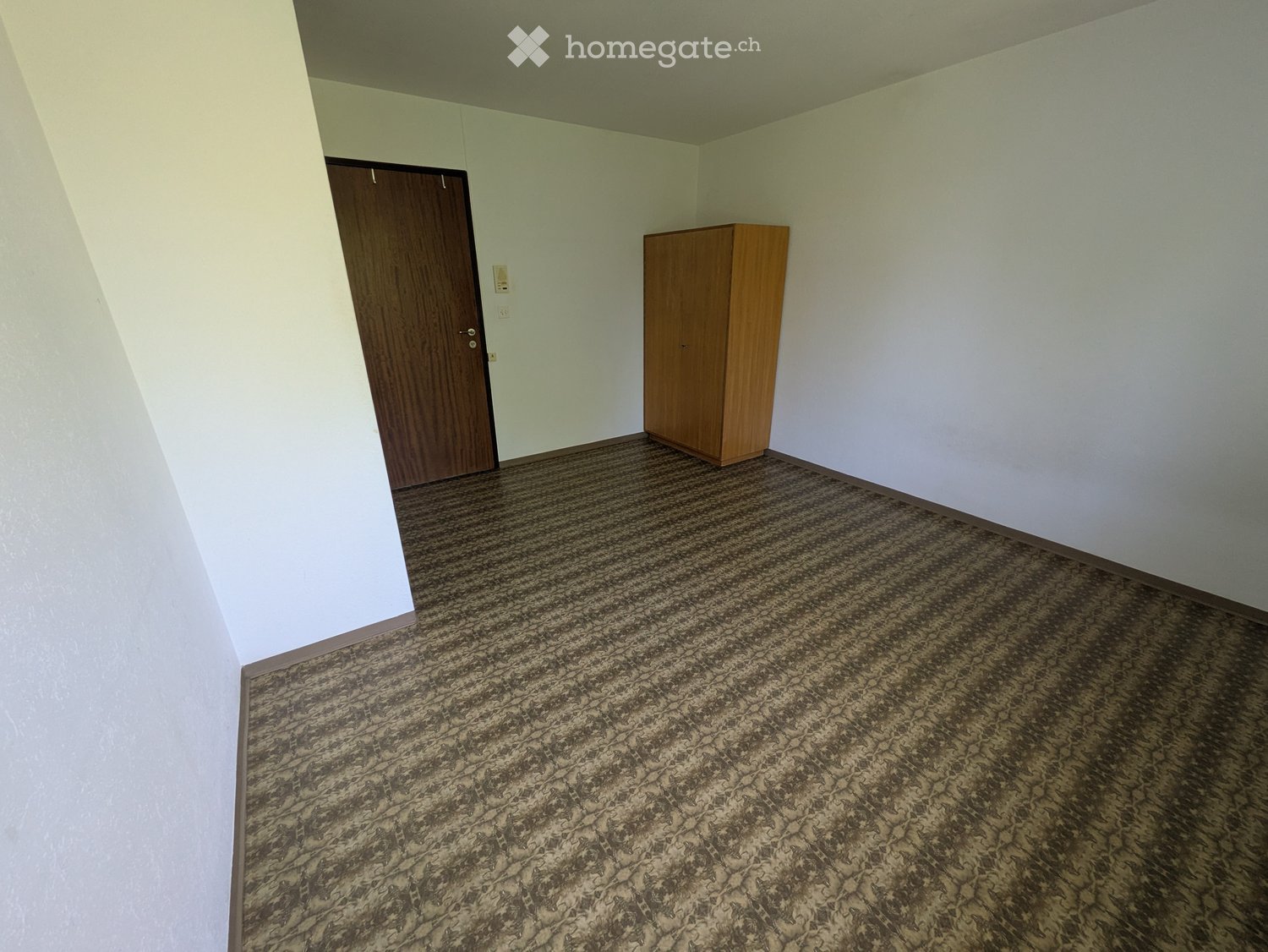 Empty room with light-colored walls, a wooden door, and a small wooden cabinet. The floor is covered with patterned carpeting.