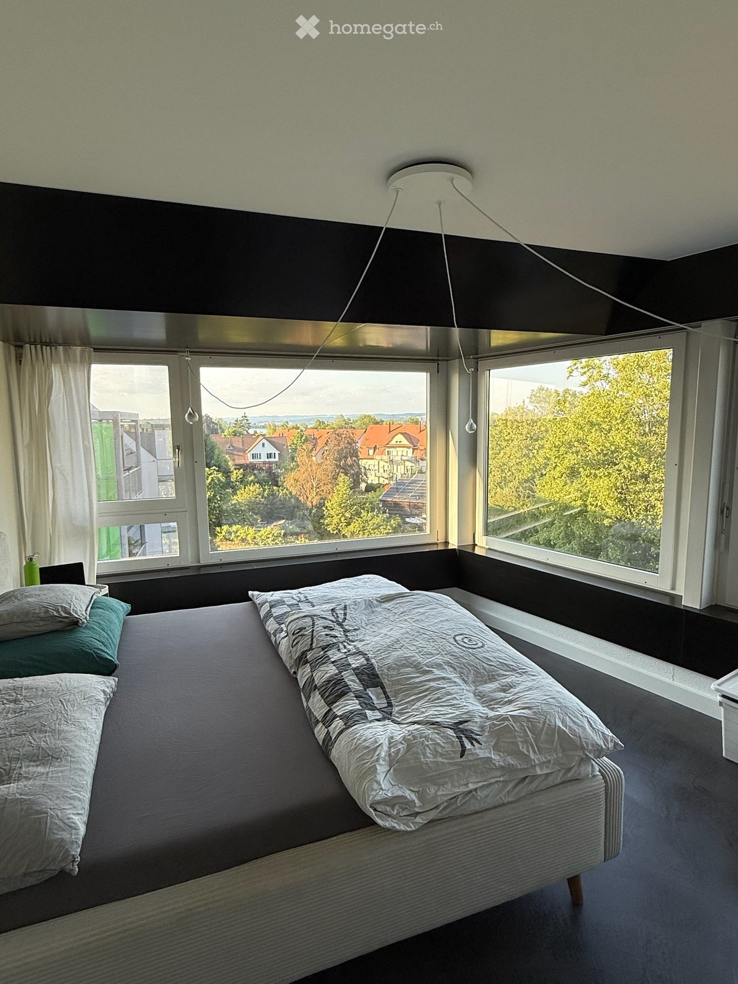 Modern bedroom with large windows offering a scenic view, featuring a neatly made bed with pillows and a duvet, a chair, and a curtain on one side.