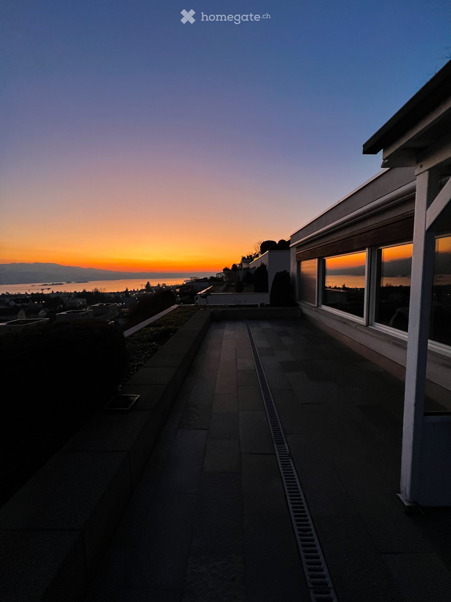 Roof terrace with stone tiles, low walls, and expansive view of the ocean and cityscape at sunset.