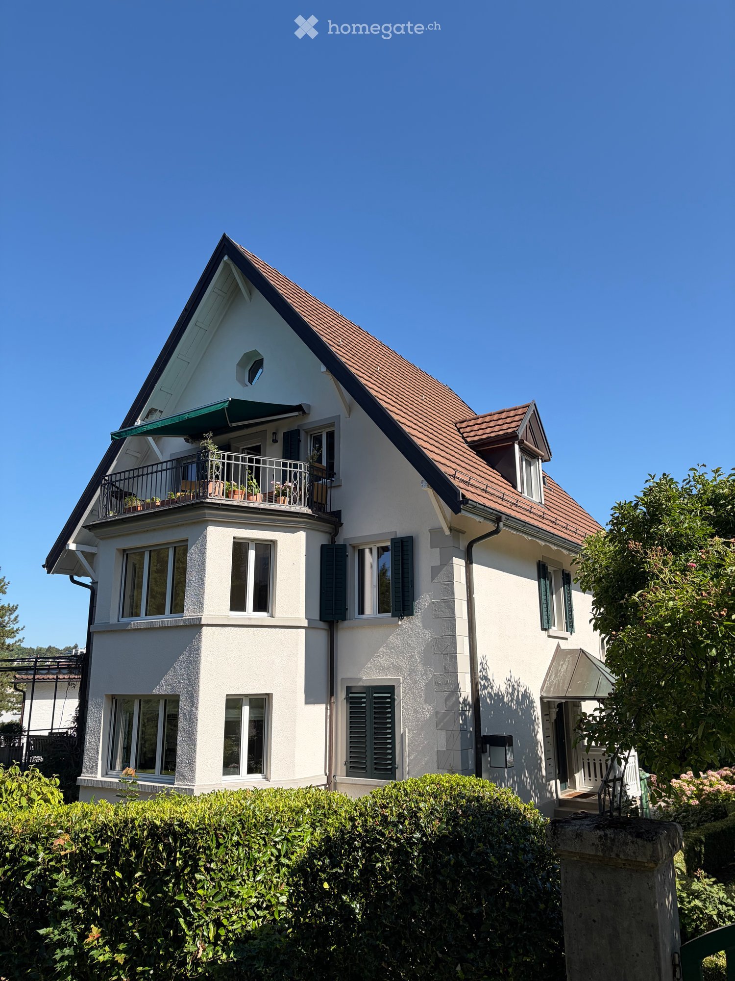 Two-story house with white exterior, brown tiled roof, multiple windows with shutters, and a small balcony with plants.