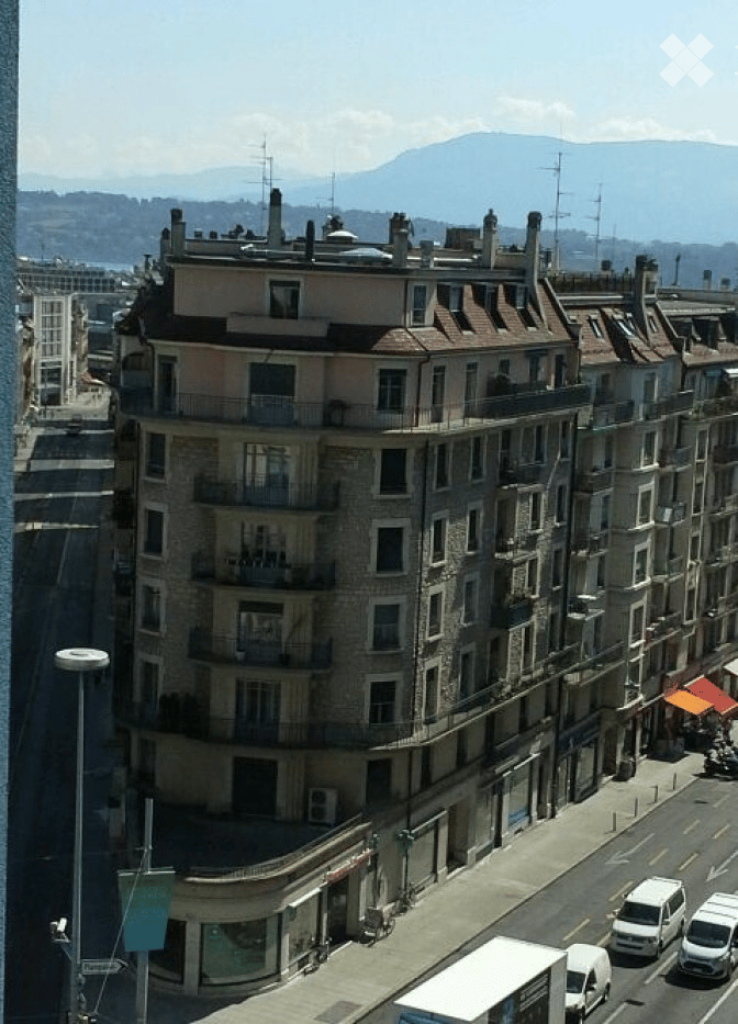 Multi-story urban building with balconies and chimneys, overlooking a city street and distant mountains.