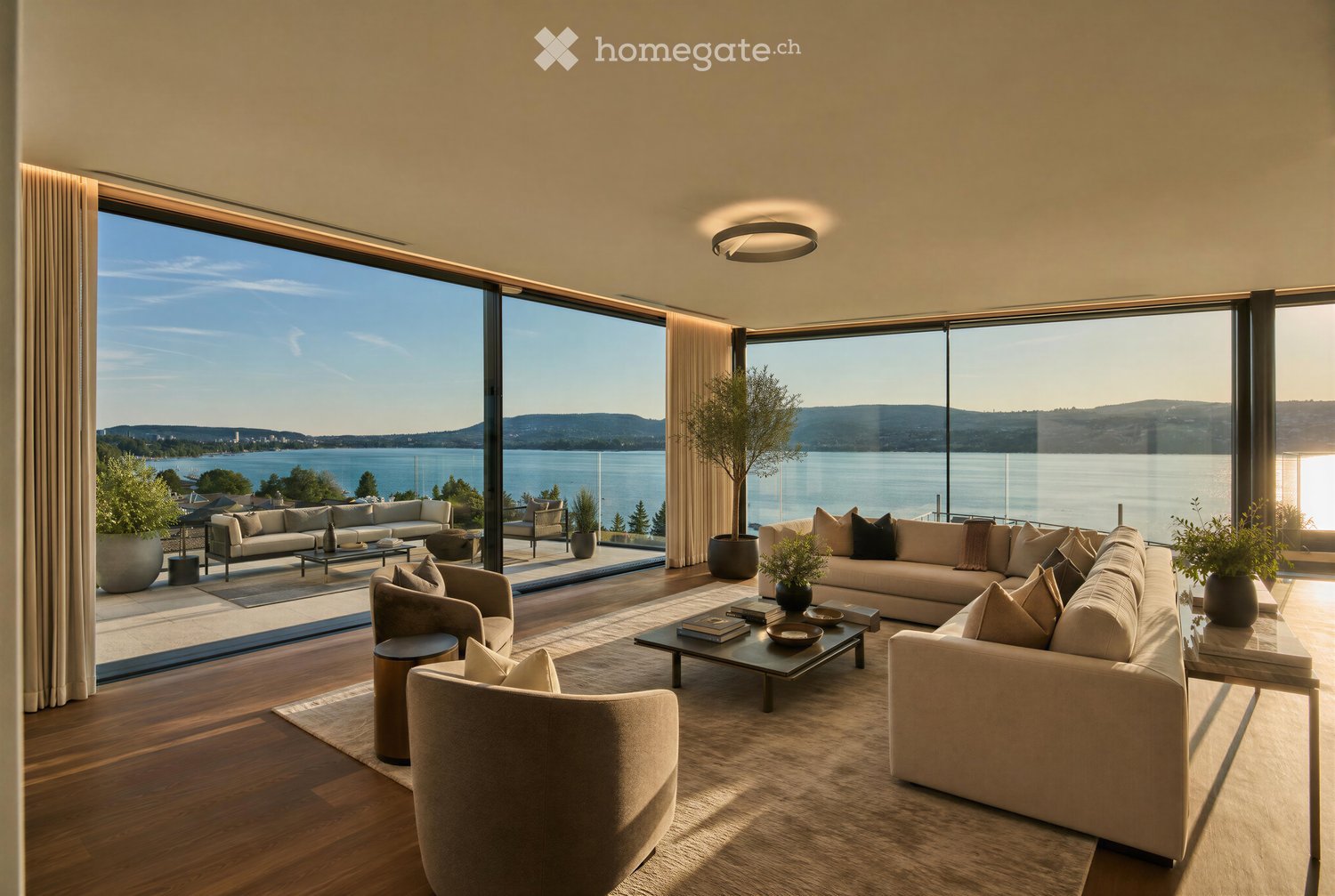 Modern living room with large windows offering a scenic lake view, featuring a beige sectional sofa, coffee table, armchairs, and decorative pillows.