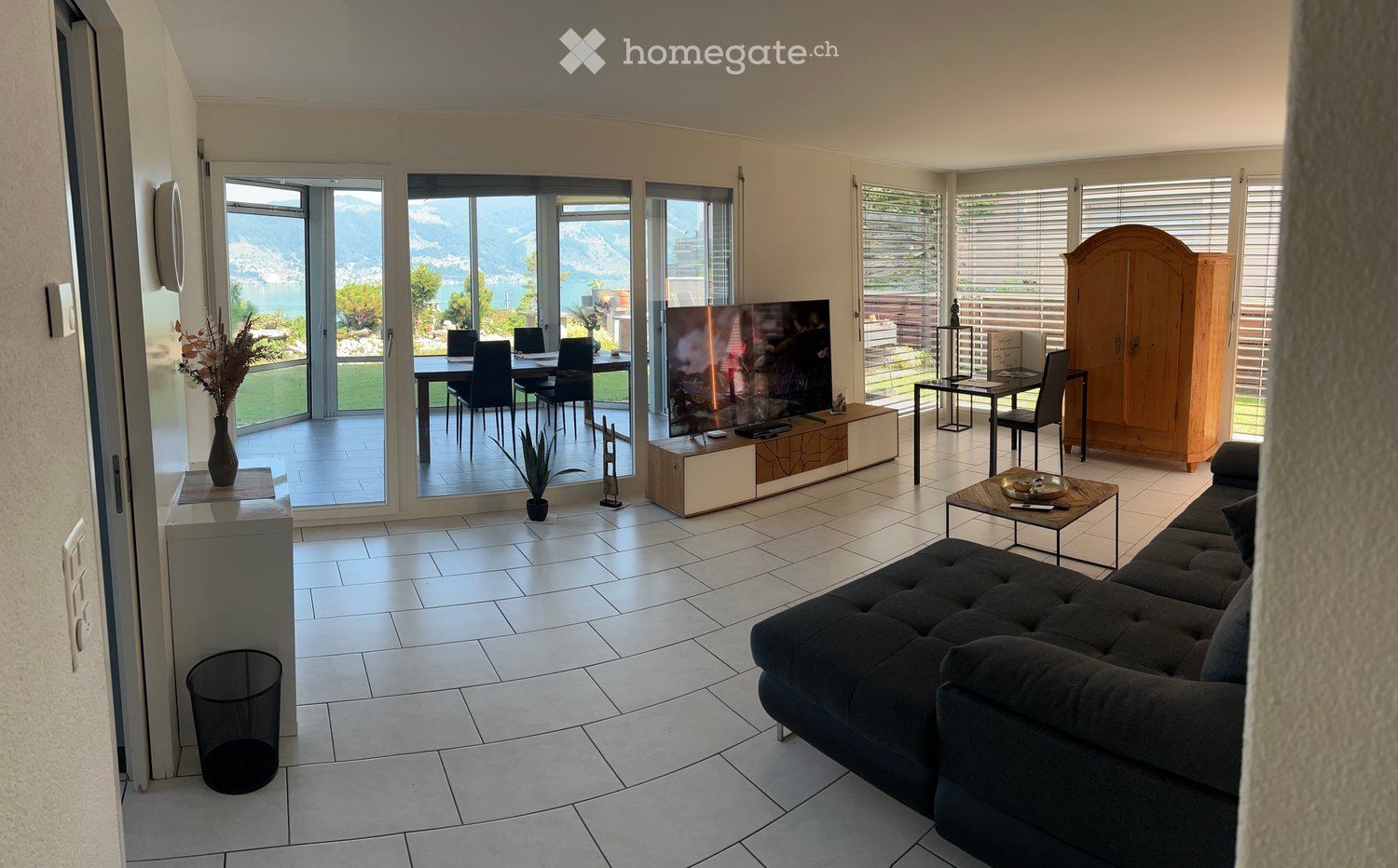 Modern living room with large windows offering a scenic view, featuring a black sofa, coffee table, TV stand, dining area with table and chairs, and a wooden cabinet.