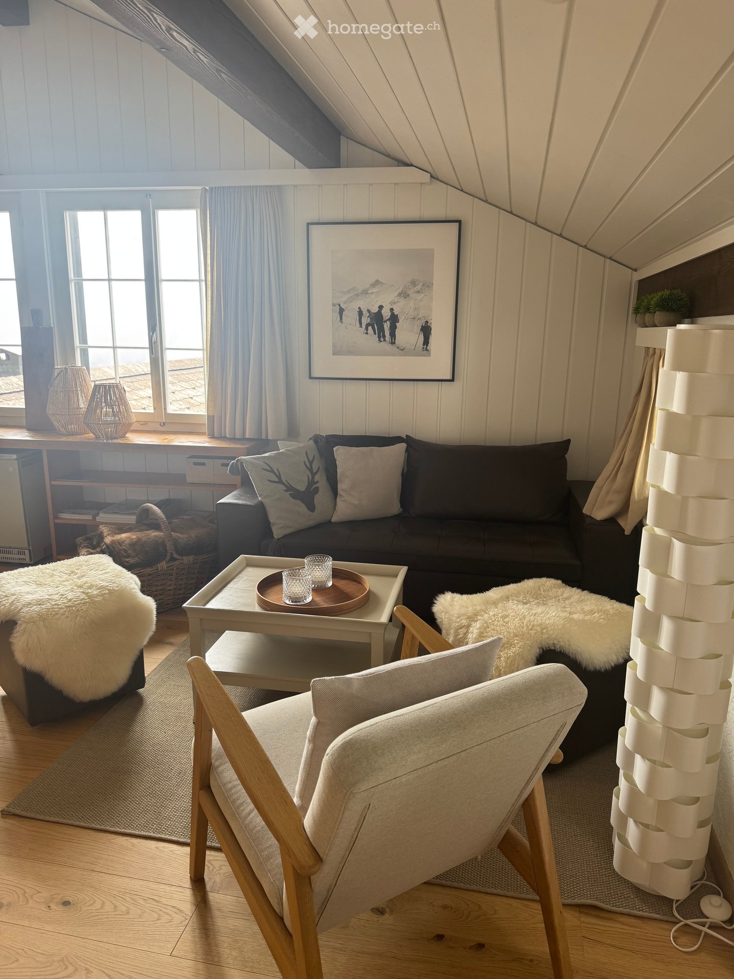 Cozy attic living room with slanted wooden ceilings, large windows, a dark grey sofa with decorative pillows, a wooden coffee table with glassware, a light grey armchair, and a fluffy white throw on a side table.