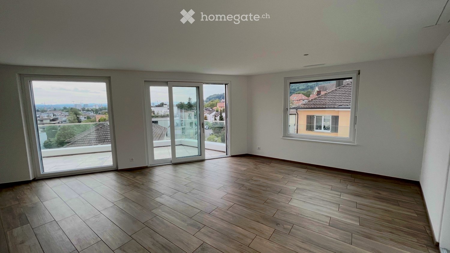 Empty room with wooden flooring, large sliding glass doors leading to a balcony, and a window overlooking a residential area.