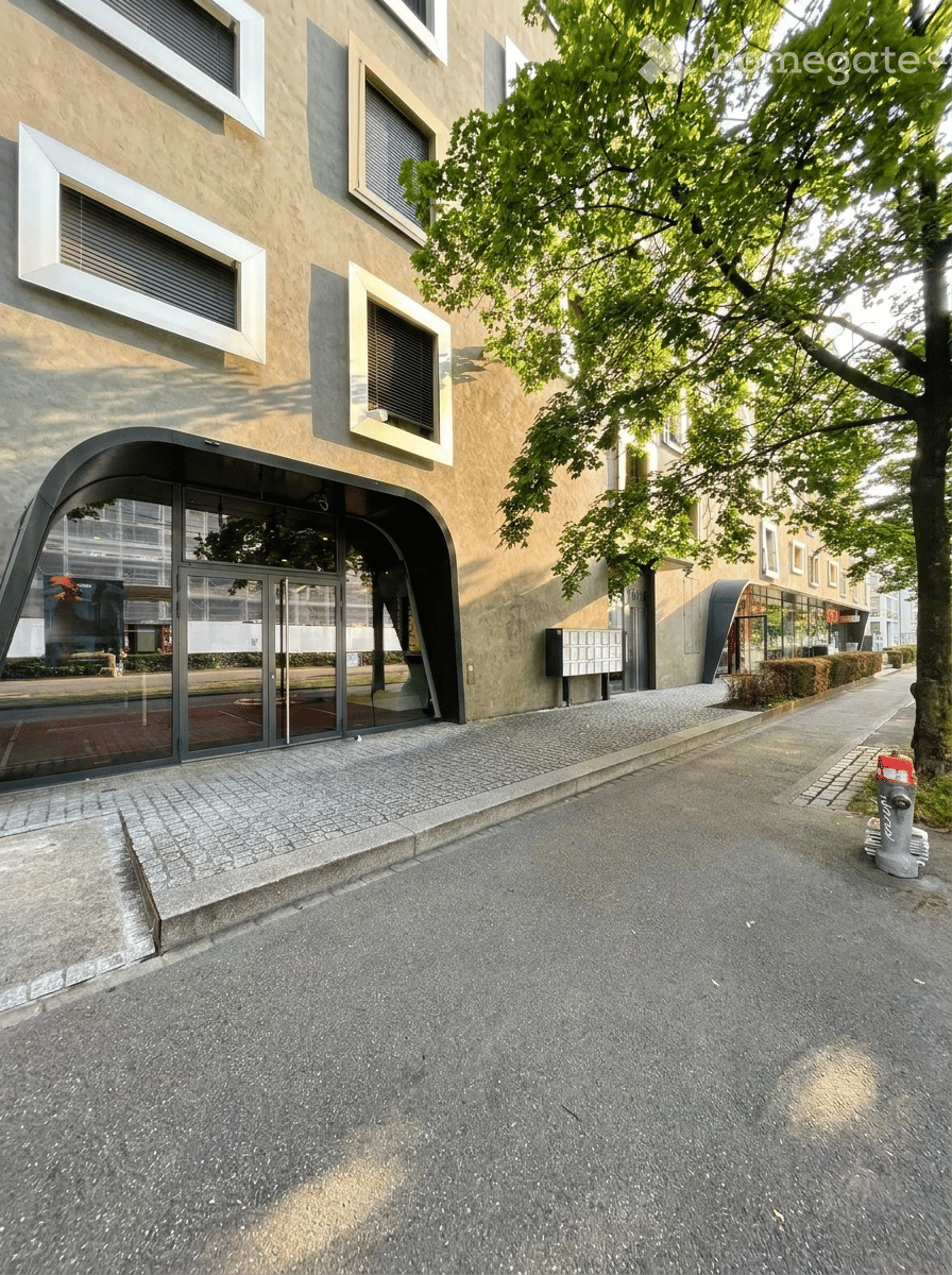 Modern building facade with large windows and a curved entrance, surrounded by trees and a paved sidewalk.