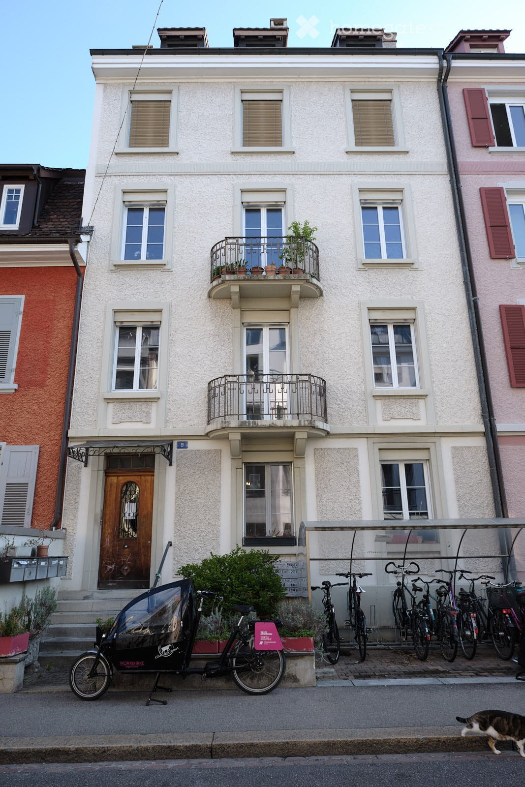 Two-story residential building with white facade, multiple windows, balconies with potted plants, and bicycles parked in front.