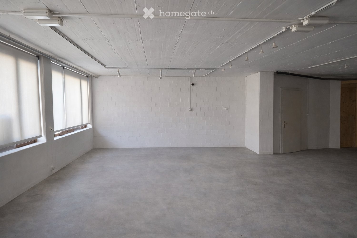 Empty room with white brick walls, large windows, and a concrete floor. No furniture or equipment is present.
