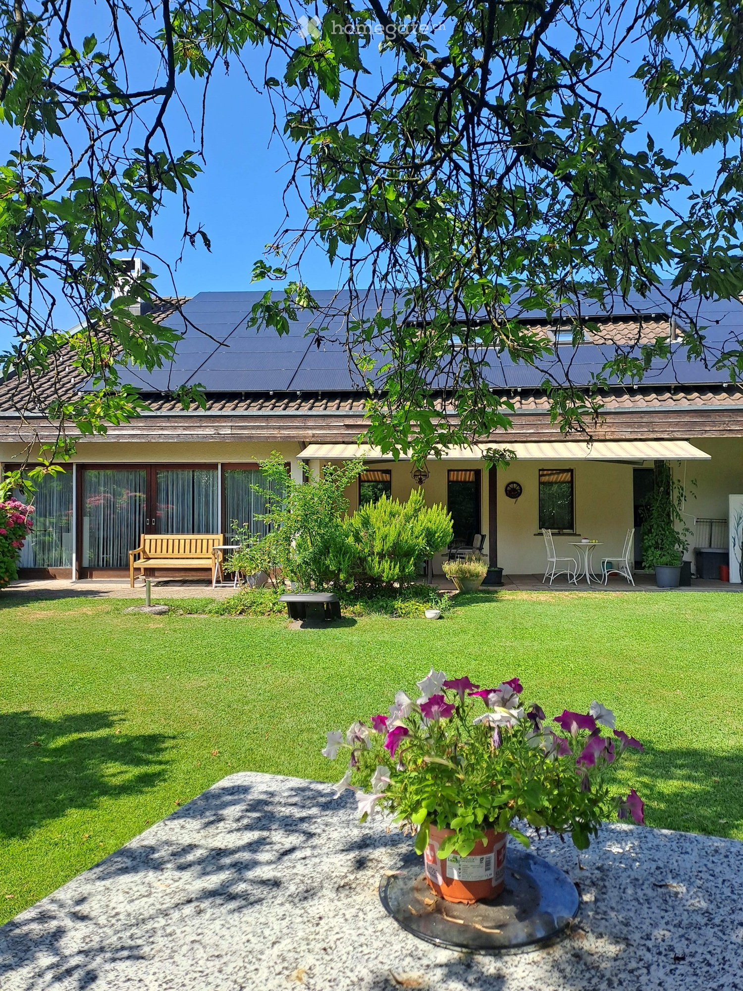 Well-maintained garden with a lawn, potted plants, a wooden bench, outdoor furniture, and a house with solar panels on the roof.