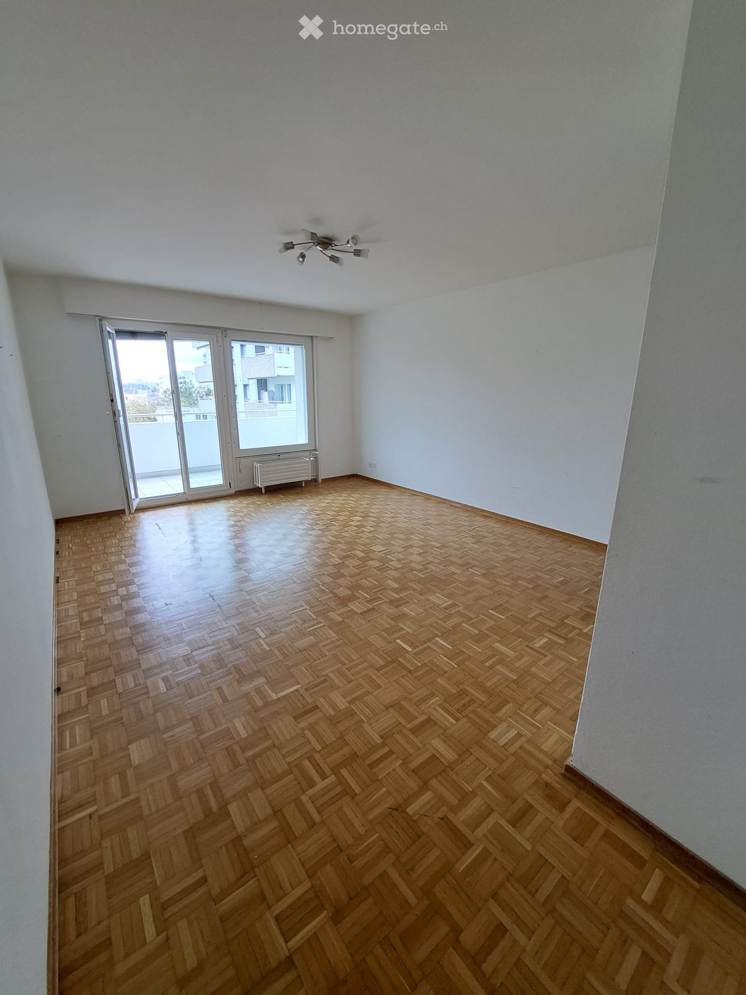 Empty room with wooden herringbone flooring, white walls, and a large sliding glass door leading to a balcony.