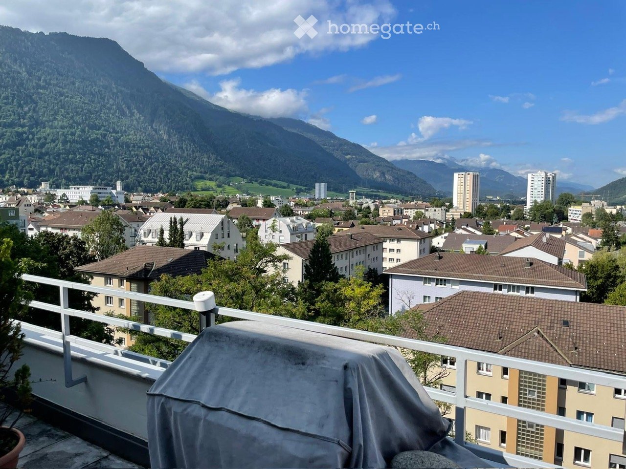 Balcony with a scenic view of a cityscape and mountains, featuring a covered object and a small potted plant.
