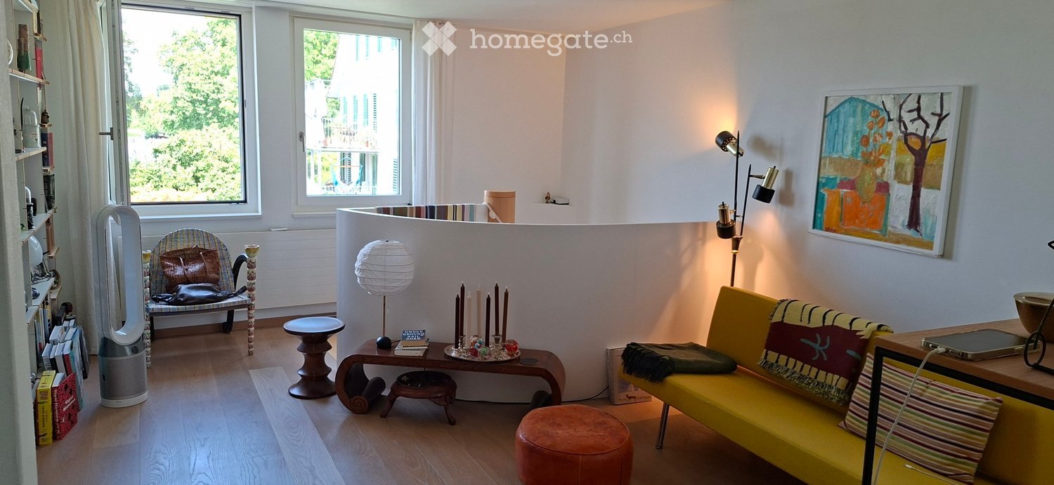 Bright living room with wooden flooring, large windows, a yellow sofa with colorful cushions, a wooden chair with a patterned cushion, a small round table, a floor lamp, and a bookshelf with books and decorative items.